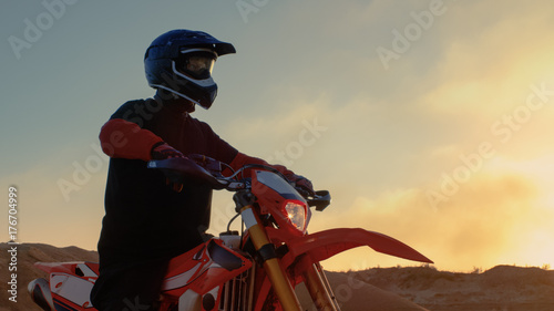 Professional FMX Motorcycle Rider Prepares to Start Driving on His Bike Over Hard Sandy Off-Road Terrain. Sun is Setting.