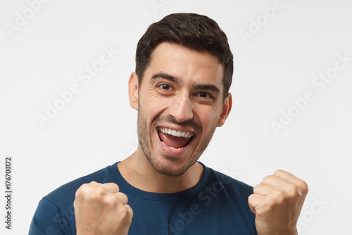 Young attractive male shouting while his team win, raised both fist in victory gesture, isolated on gray background
