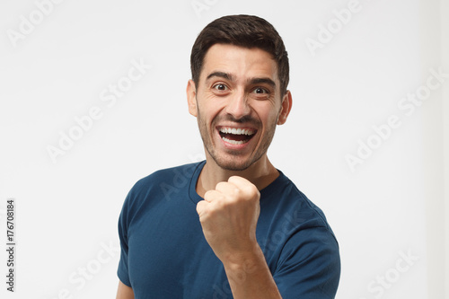 Indoor photo of good-looking male isolated on gray background celebrating victory and acting as if he is winner, squeezing fists in deep emotional expression of happiness and luck