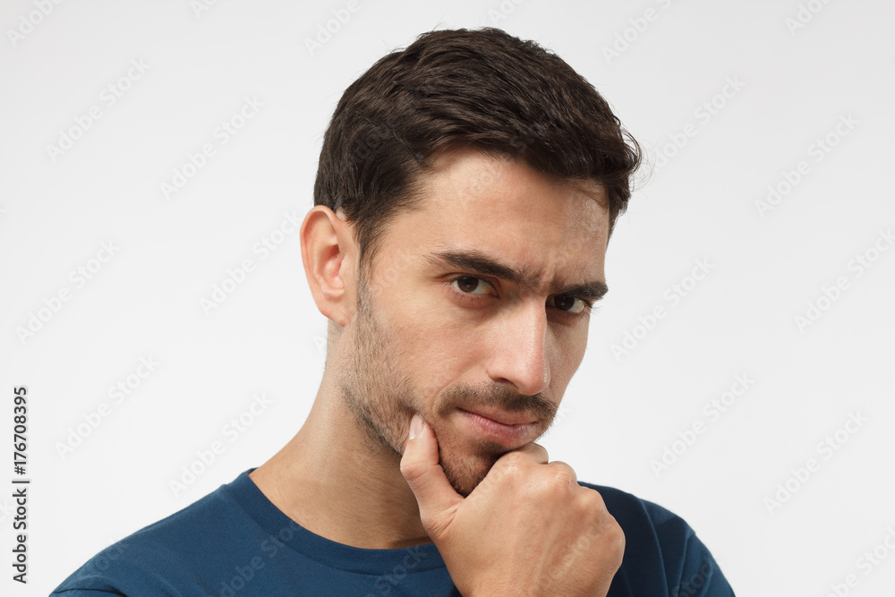 Close up portrait of young handsome male in blue t-shirt touching his chin with hand thoughtfully looking at camera. Doubt concept Close up portrait of young handsome male in blue t-shirt touching his chin with hand thoughtfully looking at camera. Doubt concept
