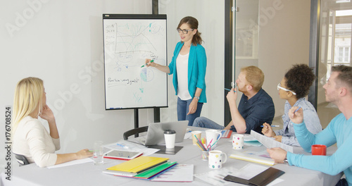 Young casual woman standing near board drawing graphics for coworkers showing statistics while working together.