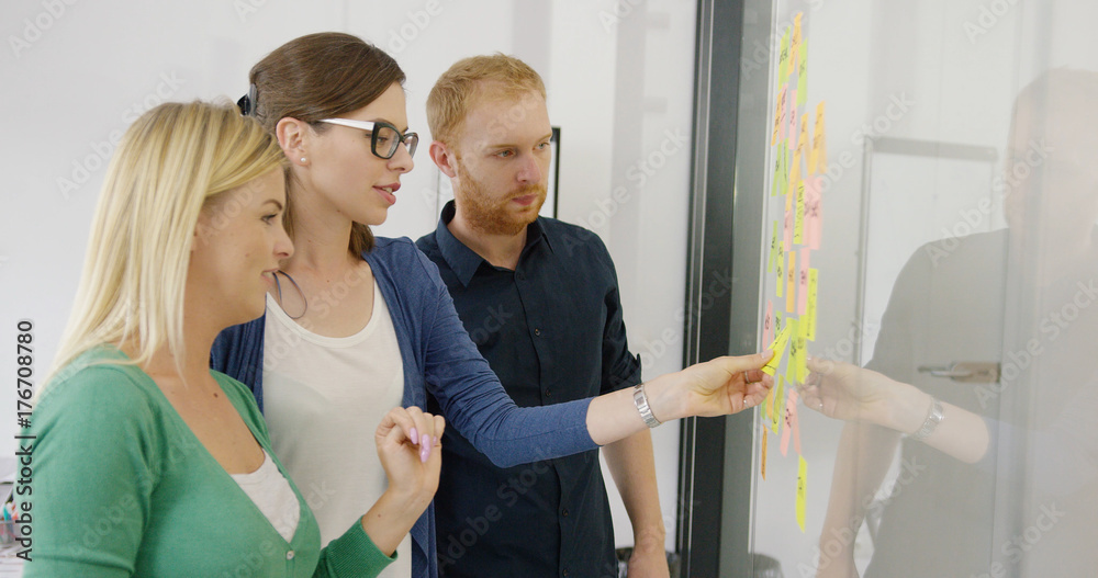 Obraz premium Side view of two women and male coworker creating new ideas on project and putting stickers on glass wall of office.