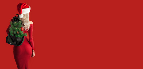 girl in a red dress on a red background in a santa hat with a backpack out of which the branches of a New Year tree stick out