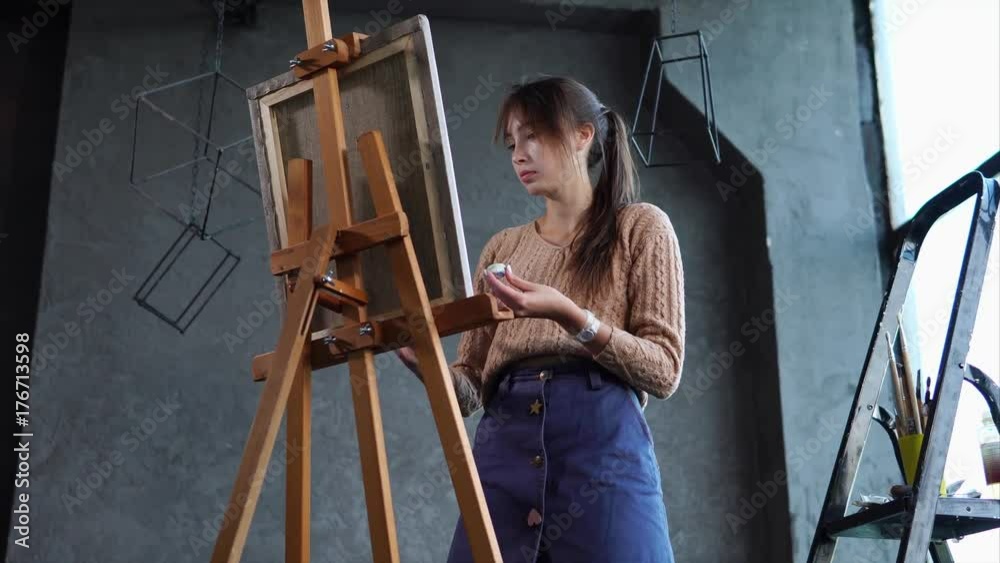 Quite a skilfull artist is drawing a picture on an easel in art studio with black walls. She is painting with a brush very fast. Quite a skilfull artist is drawing a picture on an easel in art studio with black walls. She is painting with a brush very fast.