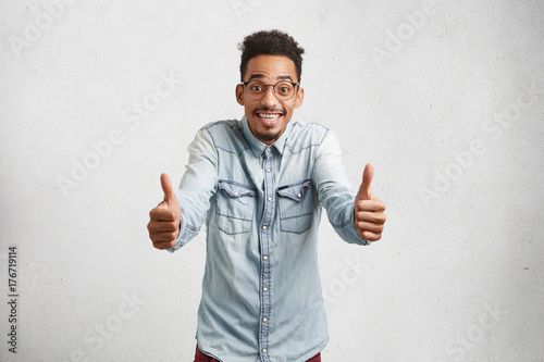 Overjoyed young male with shaggy hair, mustache, raises thumbs, shows ok sign, being delightful with results of entry exams, has broad smile, demonstrates white even perfect teeth, dressed casually. photo