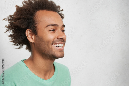 Sideways portrait of glad young smiling dark skinned male with crisp bushy hair, looks aside, being happy to notice friends and relatives who came to visit him, feels their support and great love photo