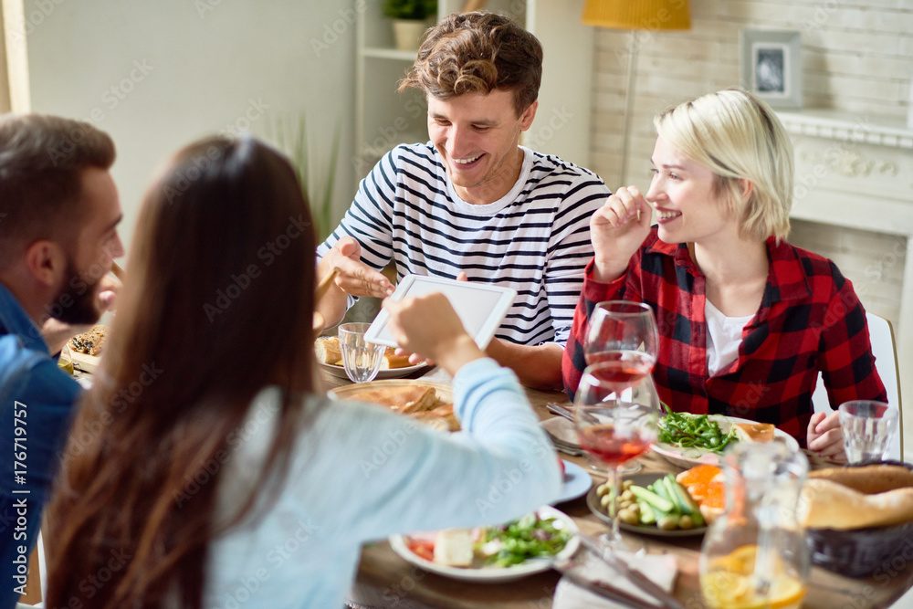 Portrait of young people laughing happily looking at digital tablet while enjoying dinner with friends sitting at big table with food