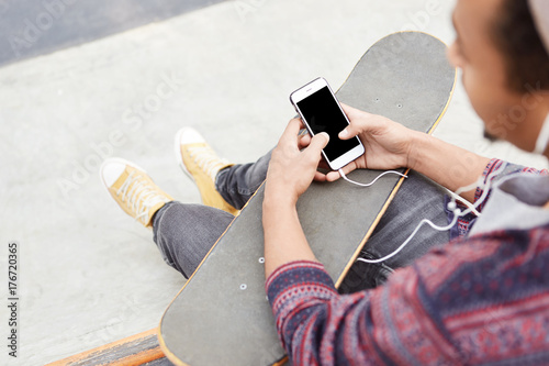 Cropped shot of male teenger has active lifestyle, skateboards on skate play, recieves message from friend, invites him to go in for extreme sport together, listens radio online with earphones photo