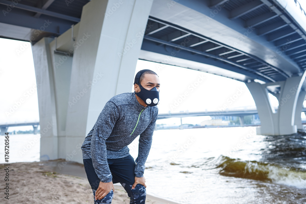 Lifestyle picture of tired young African American male runner in training mask resting, having break during evening running workout on city beach, catching his breath with hands on his knees
