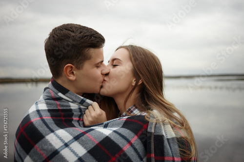 Close up portrait of cute young couple kissing and cuddling under warm plaid, spending weekend outdoors by the river, feeling happy and carefree. Love, romance, dating and togetherness concept