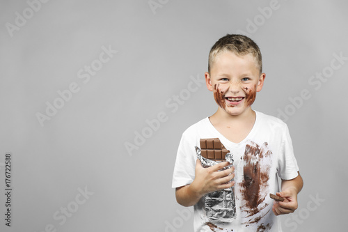 Little boy holds a bar of chocolate in his grin and has smudged clothes