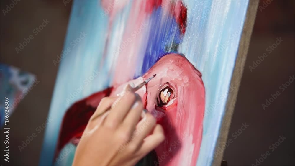 Artist is painting an eye and beak of pink flamingo by oil paints on a canvas. Close-up shot of her hand, holding paintbrush, in workshop. Artist is painting an eye and beak of pink flamingo by oil paints on a canvas. Close-up shot of her hand, holding paintbrush, in workshop.
