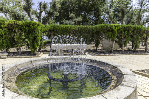 Fountain basin with bubbling up water jets with green garden trimmed hedge and stone bench on background from side