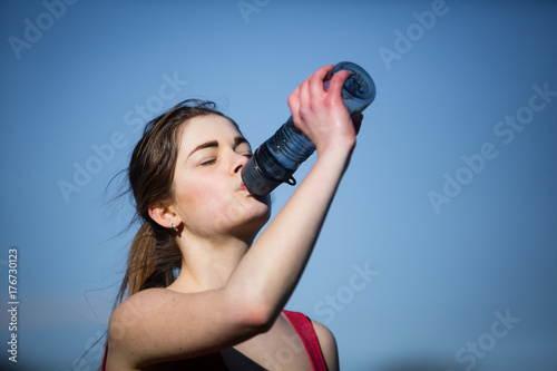 Beautiful blonde girl drinking water from a bottle after running. Shot outdoors. Blue background