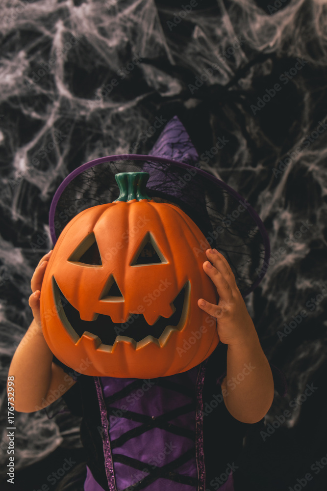 Naklejka premium Little girl dressed as a witch holding a pumpkin against a dark background with spiderwebs