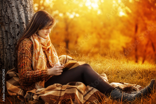 Beautiful young brunette sitting on a fallen autumn leaves in a park, reading a book