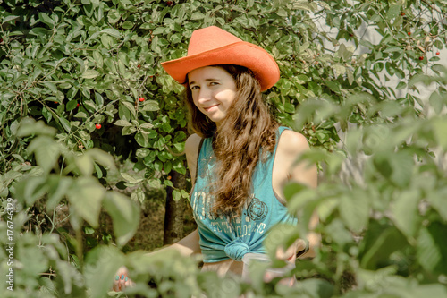 Close up portrait of curly beautiful girl with orange cowboy hat picking cherries. Woman is among the trees with berries