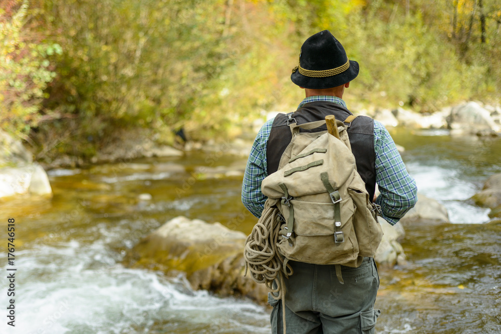 An elderly man in a hat with a backpack is on the rock of a mountain river. Senior traveler on a mountain river.
