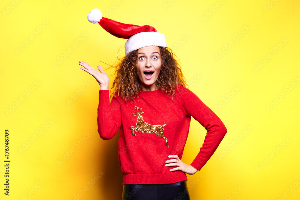 Young Santa woman in christmas hat wearing red sweater with deer smiling holding christmas gifts and having fun laughs happy rejoices touches on yellow background in the studio Young Santa woman in christmas hat wearing red sweater with deer smiling holding christmas gifts and having fun laughs happy rejoices touches on yellow background in the studio