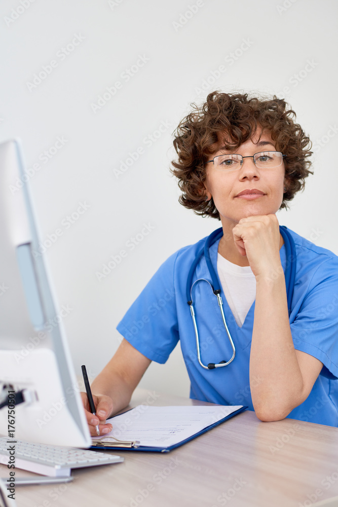 Portrait of friendly female doctor sitting at desk in office and posing, looking at camera Portrait of friendly female doctor sitting at desk in office and posing, looking at camera