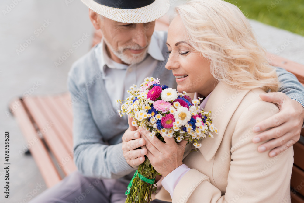 Two pensioners are sitting on a bench in the alley. The aged man gave the woman flowers. He holds her hand Two pensioners are sitting on a bench in the alley. The aged man gave the woman flowers. He holds her hand