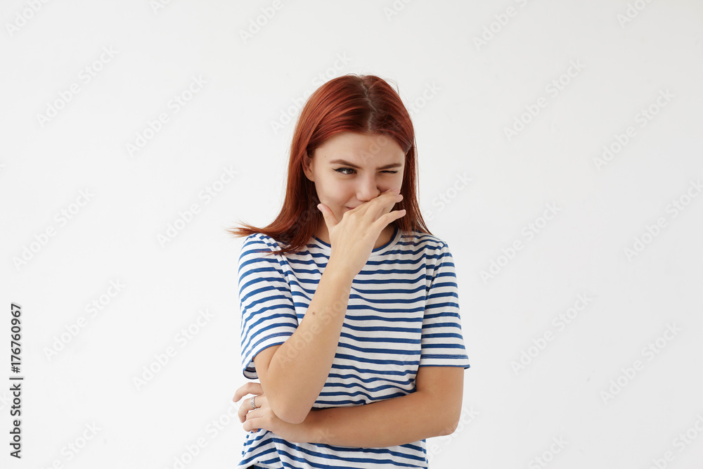 Isolated shot of funny young Caucasian woman with red colored hair covering nose with hand while sneezing. Pretty girl wearing stylish striped t-shirt resisting laughter, posing at blank studio wall Isolated shot of funny young Caucasian woman with red colored hair covering nose with hand while sneezing. Pretty girl wearing stylish striped t-shirt resisting laughter, posing at blank studio wall