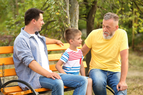 Cute boy with dad and grandfather sitting on bench in park © Africa Studio