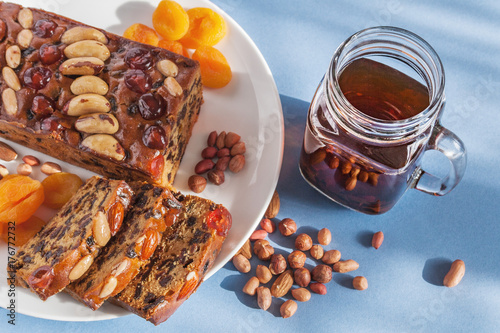 Fototapeta Naklejka Na Ścianę i Meble -  All butter cake packed with fruits and topped with glazed cherries and mixed nuts. Accompanied with a cup of black tea, dried apricots and mixed nuts. Light blue background