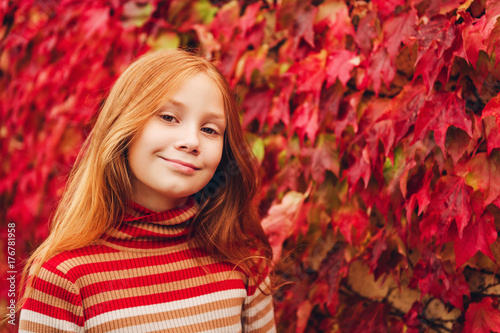 Outdoor close up portrait of adorable preteen girl of 10 years old, leaning on red ivy wall. Child with long red hair enjoying nice autumn day