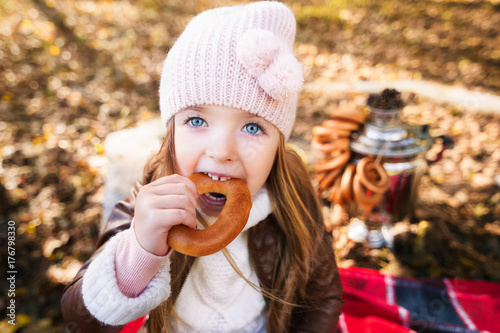A funny portrait of a little girl. The child is eating a bagel. The girl looked up in surprise. Russian samovar with bagels in the background.
