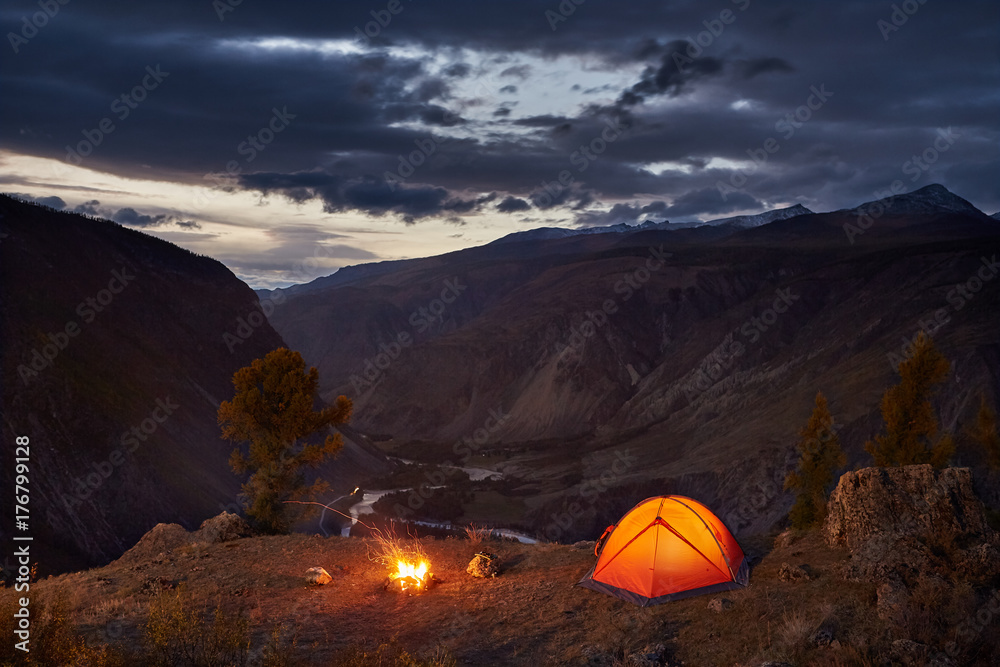 A illuminated tent and campfire in mountains in dawn