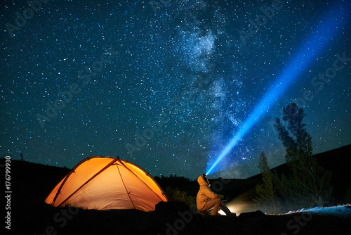 Man tourist with flashlight near his camp tent at night.