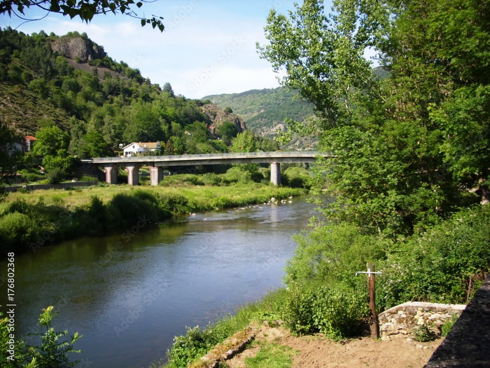 Landscape of green countryside around a road bridge spanning a river. Wooded hills and sky in the background.