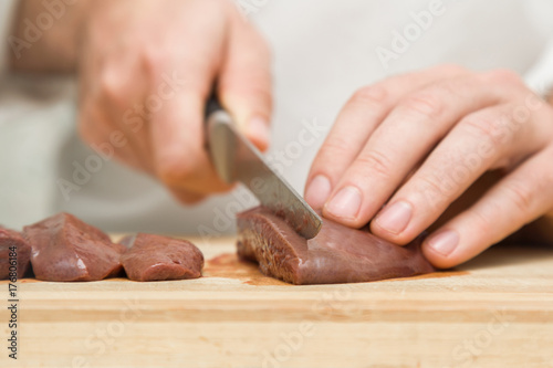 Chef's hands with a knife cutting a liver on the wooden board for pate, pancakes or cutlets in the kitchen. Preparation for cooking. Healthy food concept.