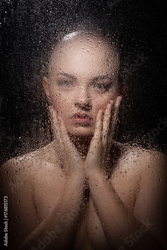 Portrait of a bald girl behind a wet glass on a black background.