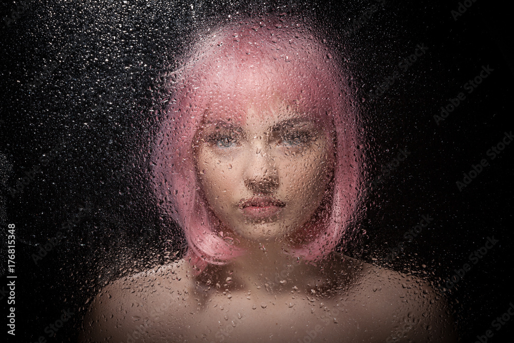 Portrait of a bald girl with pink hair behind a wet glass on a black background.