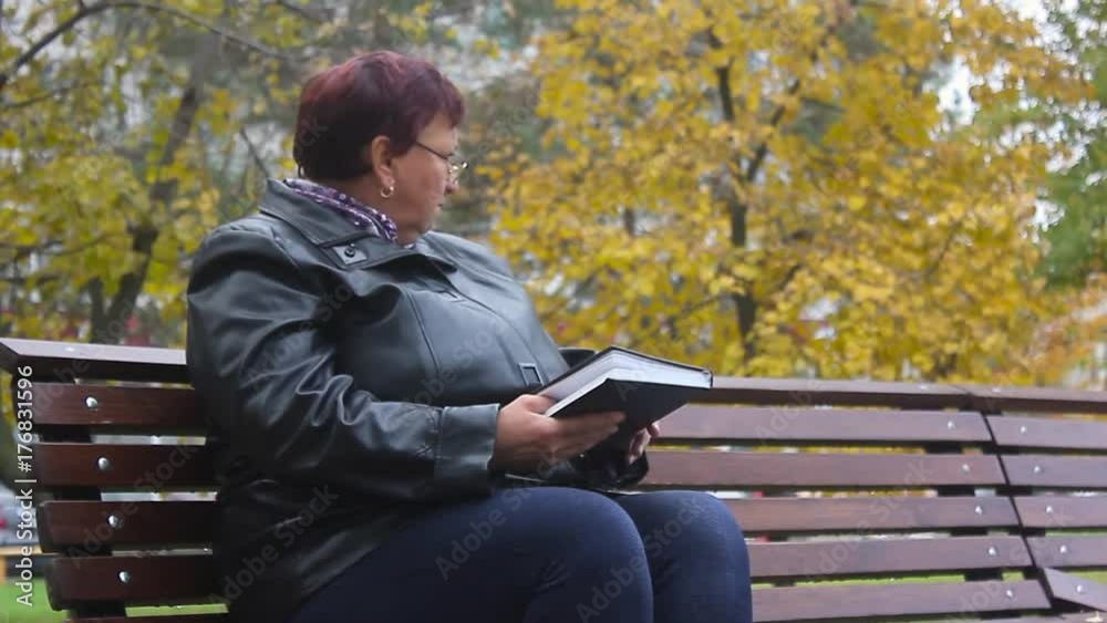 Old age, retirement and people concept - happy senior woman reading book sitting on bench at autumn park. Side view at elder woman in glasses reading book on bench in autumn park.