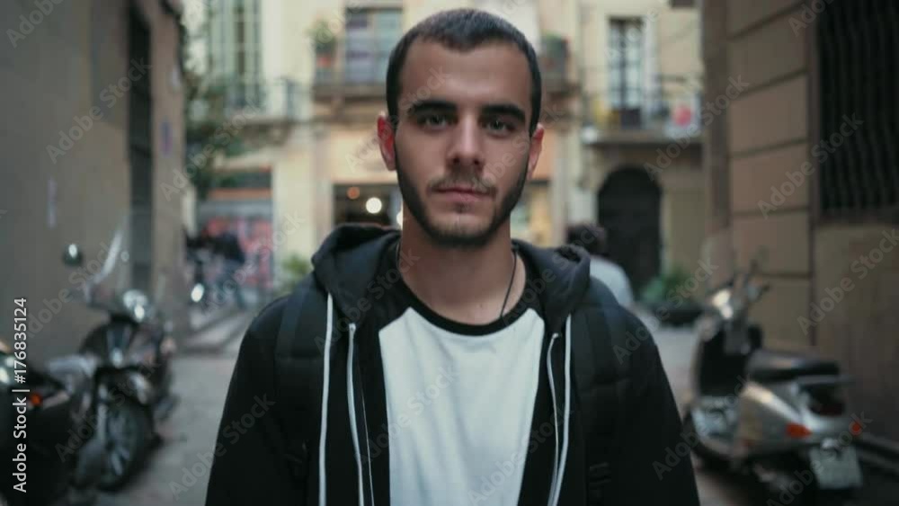 Confident young man in black urban hoodie, handsome and pleasant walks streets of european busy town or city, looks straight into camera, first serious but then gently smiles and laughs white teeth