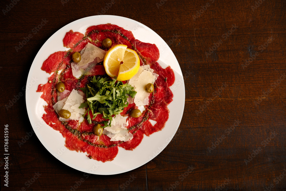Italian beef carpaccio with parmesan shavings and rocket isolated on dark brown table from above.