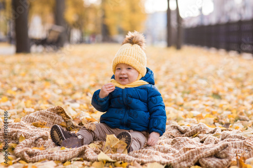  happy little boy in the sun in autumn, sitting in leaves and yellow cute hat