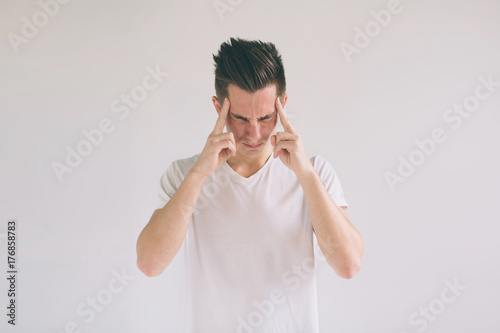 Man with glasses having troubles and headache at home. Young black-haired nerd guy wearing white t-shirt holding hands on head while found out about something horrorful. photo