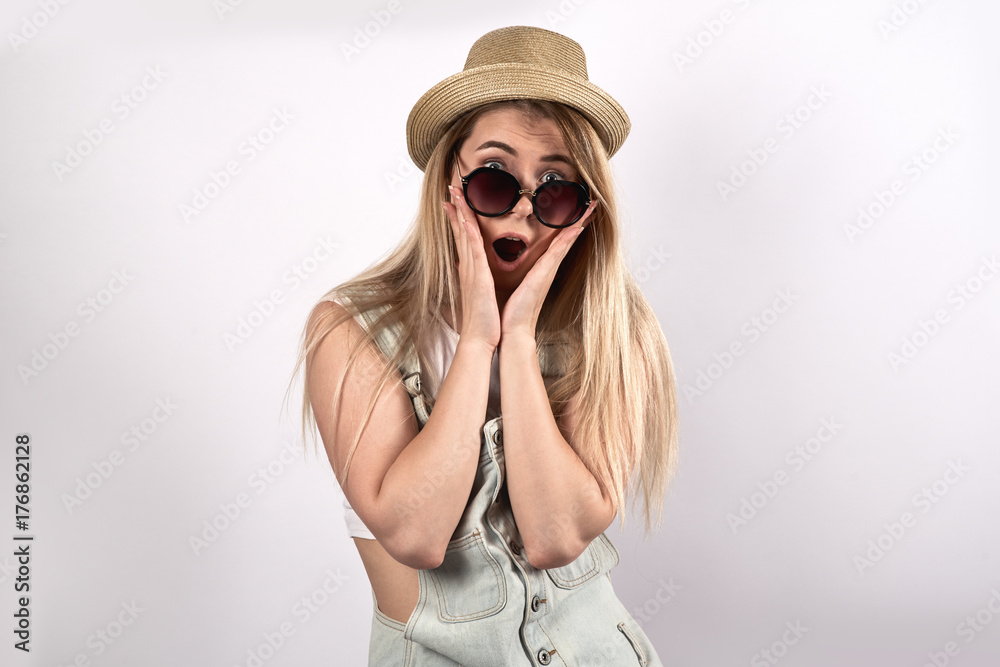 portrait of young long-haired woman in hat and gray dress. Scared shocked surprised look on her face, hands pressed to her cheeks, emotions of extreme astonishment. White background portrait of young long-haired woman in hat and gray dress. Scared shocked surprised look on her face, hands pressed to her cheeks, emotions of extreme astonishment. White background