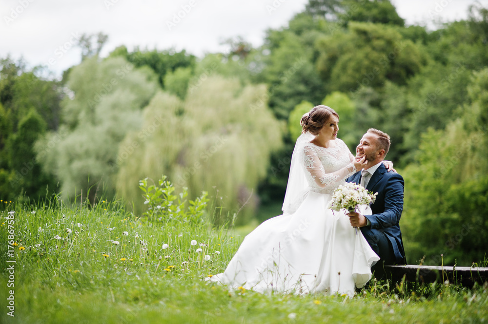 Fantastic bride sitting on a groom's lap in the meadow next to the lake on a wedding day.