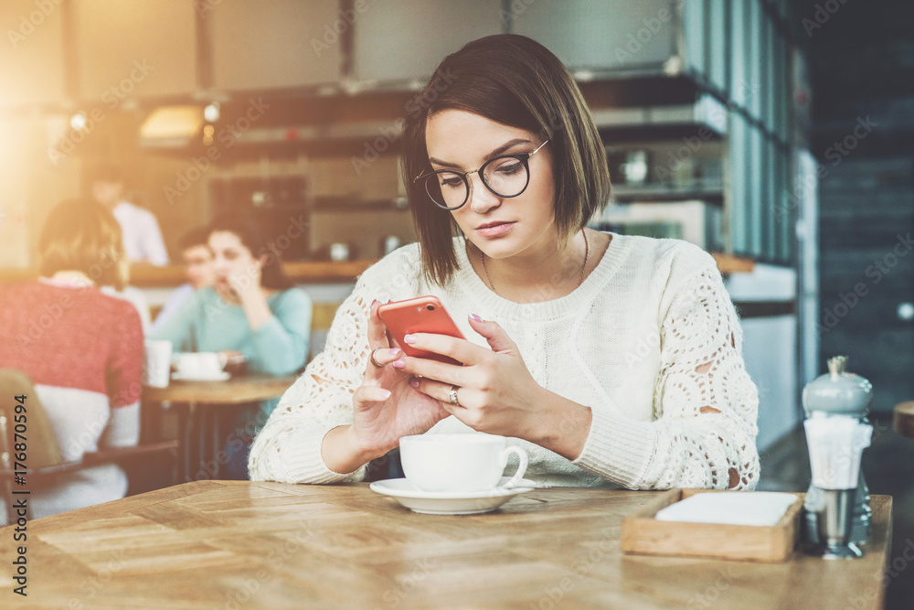 Young businesswoman is sitting in coffee shop at table and using smartphone, working. E-learning,online marketing,education.Hipster girl looking on screen of phone,checking email, blogging, chatting. Young businesswoman is sitting in coffee shop at table and using smartphone, working. E-learning,online marketing,education.Hipster girl looking on screen of phone,checking email, blogging, chatting.