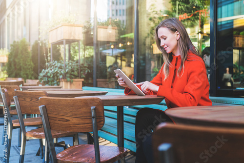 Side view. Young attractive woman in orange coat is sitting outside in cafe at table and uses tablet computer. Girl checking email, blogging, chatting, reading e-book. Social media, e-learning.