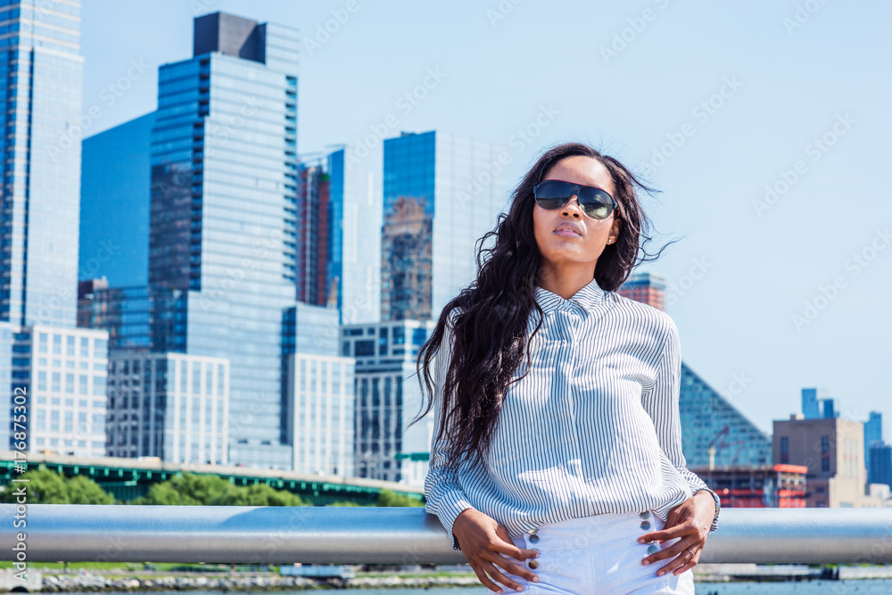 Fototapeta premium Young American Woman with long hair traveling in New York in summer, wearing white striped shirt, sunglasses, standing in business district with high buildings in Manhattan under sun, looking up..