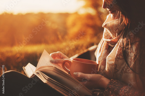 a woman sits near a tree in an autumn park and holds a book and a cup with a hot drink in her hands. Girl reading a book photo