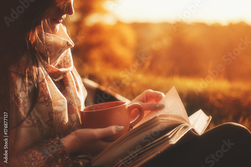 a woman sits near a tree in an autumn park and holds a book and a cup with a hot drink in her hands. Girl reading a book