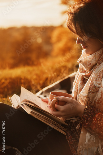 a woman sits near a tree in an autumn park and holds a book and a cup with a hot drink in her hands. Girl reading a book