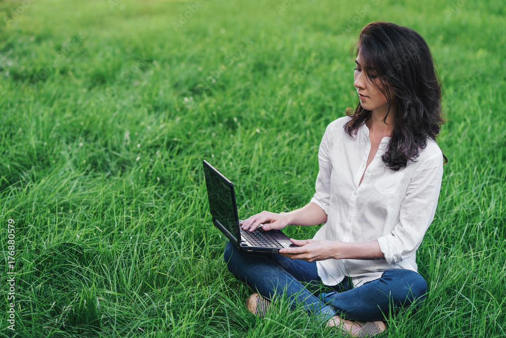 Young woman is sitting on the green grass in park with laptop. Freelance working concept. Distant learning concept. Copy space for text. Young woman is sitting on the green grass in park with laptop. Freelance working concept. Distant learning concept. Copy space for text.